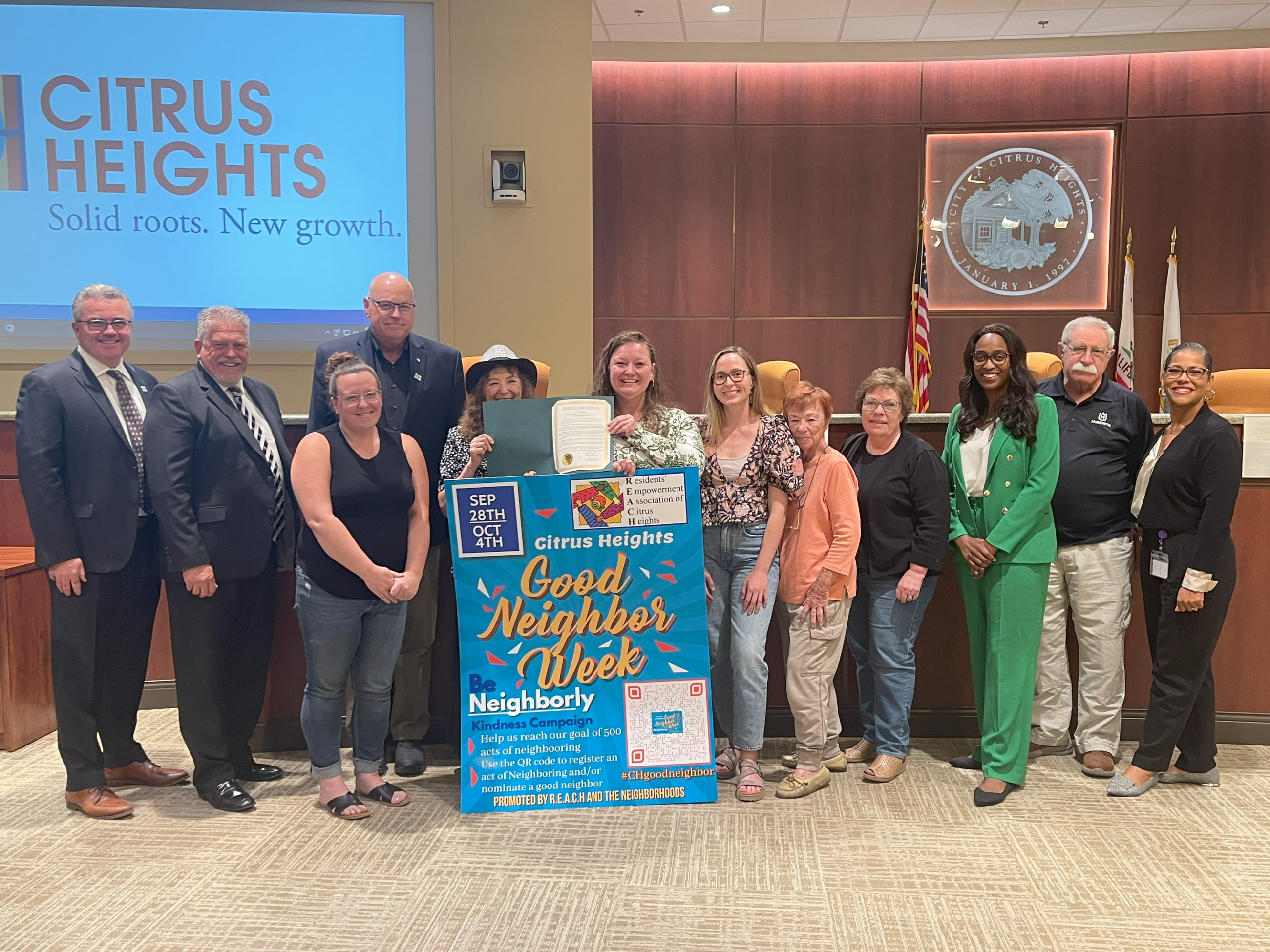 A group of men and women stand in Council Chambers with a poster in front of them.
